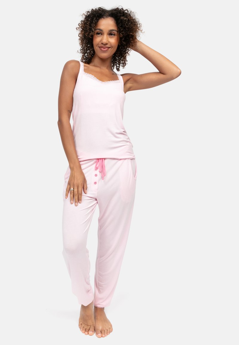 Kobieta o kręconych włosach, ubrana w jasnoróżowy bezrękawnik i pasujące piżamowe spodnie, stojąca boso z ręką na głowie.