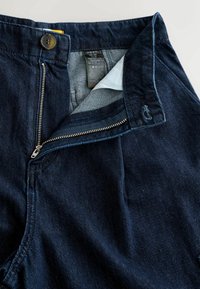 Jean en denim bleu foncé avec une fermeture éclair ouverte, bouton fermé, et une étiquette intérieure visible indiquant la marque et les instructions d'entretien.