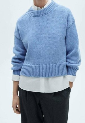 Personne portant un pull en tricot bleu clair par-dessus une chemise blanche à col, avec un pantalon noir, debout devant un fond clair uni.