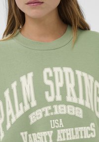 Jeune personne portant un sweatshirt vert clair avec le texte blanc "PALM SPRINGS EST. 1982 USA VARSITY ATHLETICS" sur le devant.
