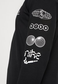 Černé tričko s dlouhými rukávy a bílými grafickými potisky zobrazujícími globální ikony a slovo "nike" v různých stylizacích písma na rukávu.