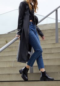 Manteau en cuir noir, haut noir ajusté, jean bleu taille haute avec hem effiloché, et bottines noires brillantes à semelles chunky, descendant des escaliers.