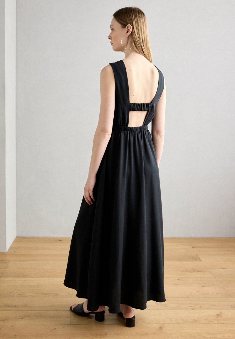 Svart maxikjole med tettsittende bodice, brede stropper og elastisk bak. Har en nøkkelhullutskjæring og en flytende skjørt. Glatt stofftekstur.