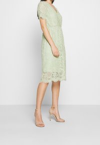 Robe en dentelle vert clair avec manches courtes, encolure en V et ourlet festonné. Le tissu présente des motifs floraux et des textures délicates.