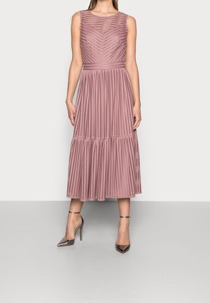 Femme portant une robe midi sans manches rose poudré avec des rayures verticales texturées et une jupe plissée, associée à des escarpins métalliques pointus.