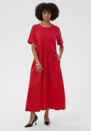 Žena s kučeravými vlasmi v jasne červenom midi šatách s krátkym rukávom a vreckami, obutá v čiernych vysokých podpätkoch, stojaca pred jednofarebným bielym pozadím.