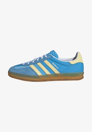 Zapatilla Adidas Gazelle azul con rayas crema, suela de goma, cordones blancos y lengüeta trasera crema, mostrada de perfil lateral mirando hacia la derecha.