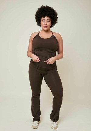 Donna con capelli ricci che indossa una canotta marrone scuro aderente e pantaloni, si sistema la cintura, in piedi davanti a uno sfondo chiaro e uniforme.
