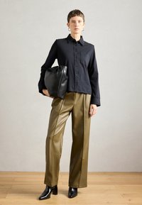 Črna srajca z gumbi in ovratnikom ter žepom na prsih, v kombinaciji z olivno zelenimi hlačami z visokim pasom in širokimi nogavicami iz umetnega usnja ter črnimi čevlji na peto.