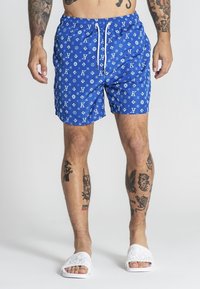 Blå badshorts med ett upprepande vitt logomönster, snörmidja och avslappnad passform, i kombination med vita slidesandaler.