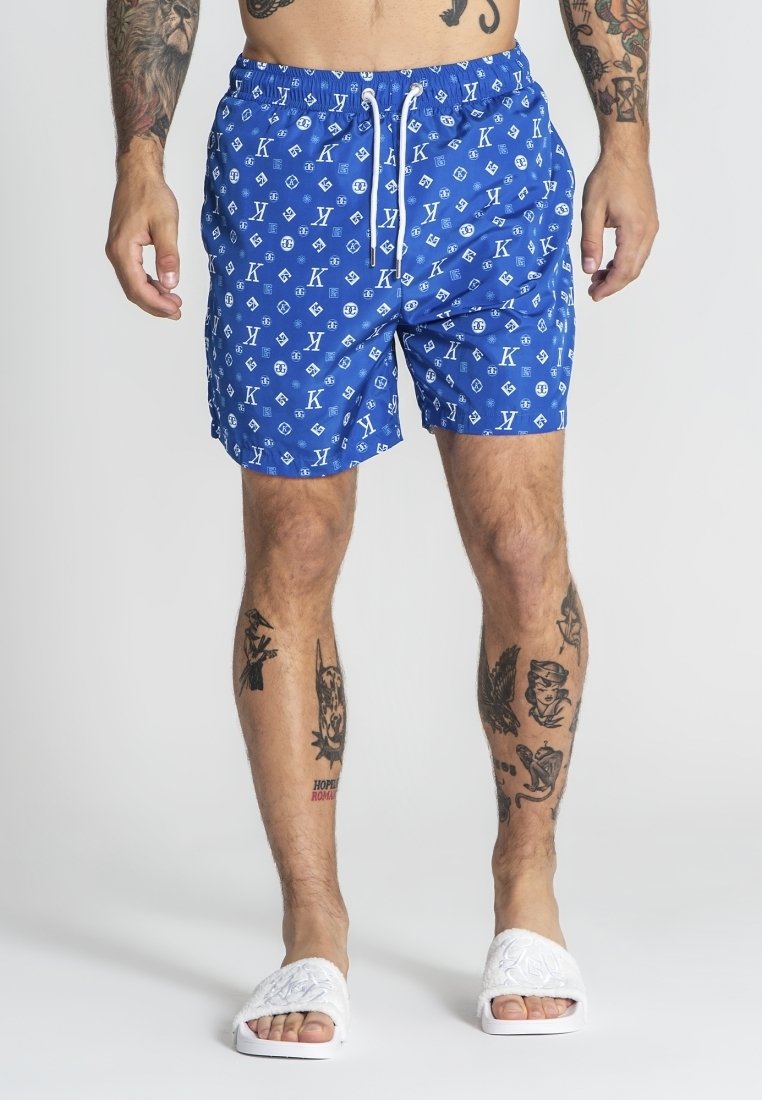Blå badshorts med ett upprepande vitt logomönster, snörmidja och avslappnad passform, i kombination med vita slidesandaler.