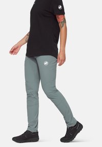 Pantalons de randonnée légers et gris, au design ajusté, avec des panneaux aux genoux et un logo sur la hanche, associés à des chaussures de sport noires.