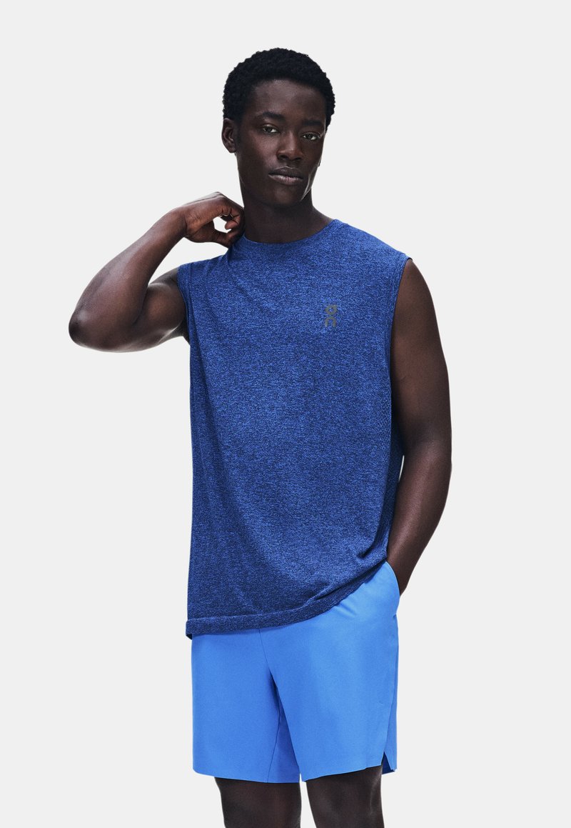 Homme portant un débardeur de sport bleu et un short bleu clair, debout avec une main derrière la nuque et l'autre dans la poche.