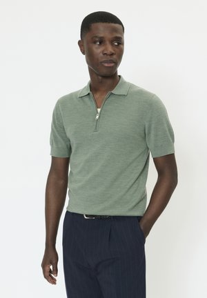 Hombre joven vistiendo un polo manga corta verde claro con cremallera frontal y pantalones azul oscuro con rayas finas, de pie con una mano en el bolsillo.