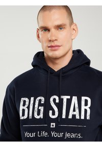 Jauns vīrietis ar zilām acīm valkā tumši zilu sporta krekli ar kapuci, uz kura rakstīts "BIG STAR Your Life. Your Jeans." baltā krāsā.