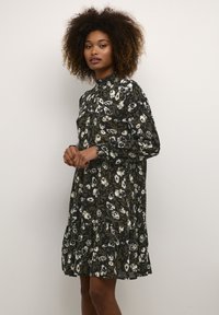 Blumenkleid in dunklem Grün mit cremefarbenen Blumen, mit hohem Kragen, langen Ärmeln und einem gestuften, rüschigen Saum. Leichtes Material.