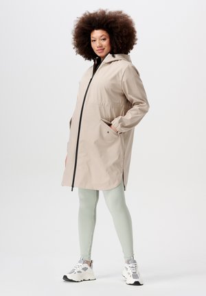 Beige wasserdichter Parka mit Reißverschluss, Kapuze, elastischen Bündchen und Seitentaschen, getragen über hellgrünen gerippten Leggings und weißen Turnschuhen.