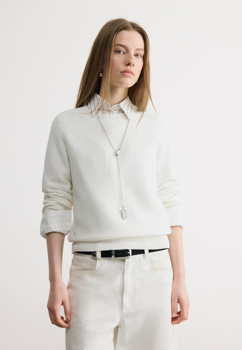 Jeune femme aux cheveux bruns lisses portant un pull blanc sur une chemise rayée à col et un pantalon blanc avec une ceinture noire et un collier argenté.