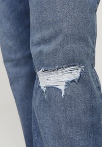 Plave traperice s istrošenim koljenom koje sadrži iščupanu rupu i svijetloplave izložene niti, tipične za ležernu odjeću.