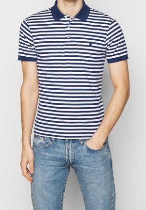Granatowa i biała polo w paski z żebrowanym kołnierzykiem i rękawami; zawiera małe logo na piersi; noszona z niebieskimi dżinsami.