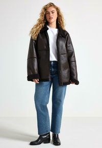 Brun læderjakke med sort shearling-kant, lynlås foran, løs pasform, båret over en hvid skjorte og blå jeans, parret med sorte ankelstøvler.