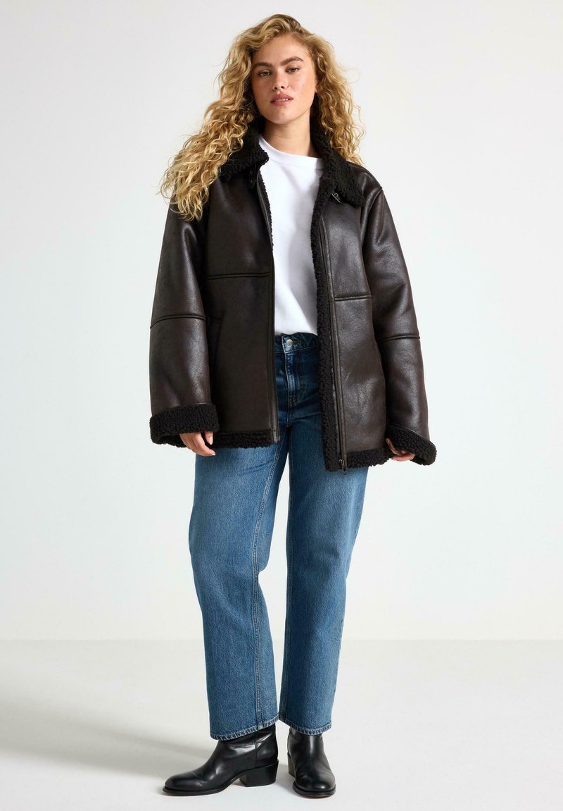 Brun læderjakke med sort shearling-kant, lynlås foran, løs pasform, båret over en hvid skjorte og blå jeans, parret med sorte ankelstøvler.