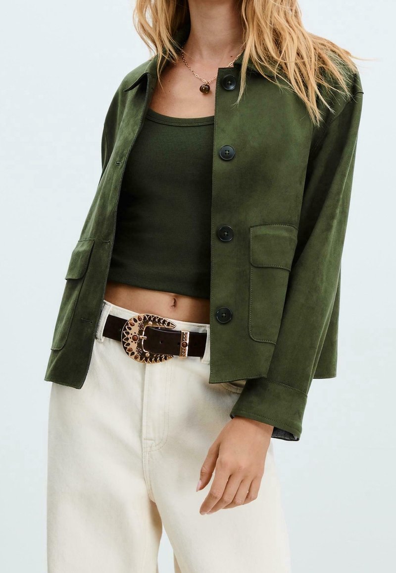 Veste en daim vert avec col, fermeture par boutons et poches avant ; associée à un débardeur olive et un jean crème taille haute.