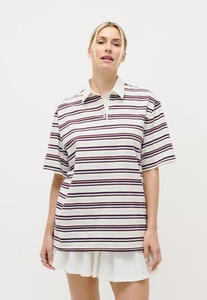 Plavolaska ženska, ki nosi ohlapno belo polo majico z bordo rdečimi horizontalnimi črtami in belo plise krilo na običajni bel ozadju.