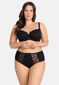 Curvy woman with long wavy hair wearing a black lace bra and matching high-waisted lace panties against a plain background.