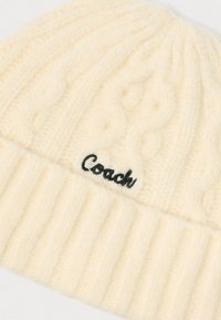 Biely pletený čiapka s kabelkovým vzorom a rebrovaným lemom. Na leme je vyšívané čierne logo "Coach". Mäkko, textúrovaná látka.