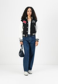 Giacca varsity in bianco e nero con toppe ricamate, abbinata a jeans blu a vita alta. La modella tiene in mano una piccola borsa scura e indossa scarpe bianche.