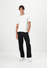 Maglietta in cotone bianco con texture, colletto a righe bianco e nero, abbinata a pantaloni neri larghi e scarpe da ginnastica bianche.