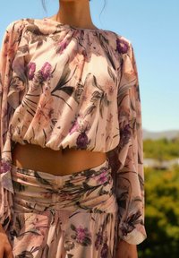 Blomstret bluse i lys pink stof med elastisk kant, med lange ærmer og foldede detaljer, parret med en matchende nederdel.