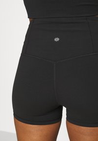 Pantaloni corto neri a vita alta in un materiale liscio e elasticizzato. Presentano un piccolo logo bianco sul retro con dettagli di cucitura a contrasto.