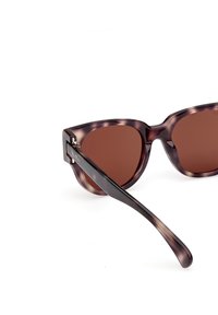 Brown-tinted sunglasses with a marbled dark purple frame. Features a unique cat-eye shape and slender arms, displaying a logo on the side.