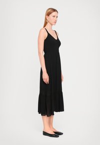 Svart maxi kjole med en tettsittende, strukturert bodice og en flytende, lagdelt skjørt. Kjolen har tynne stropper og er stylet med svarte leiligheter.