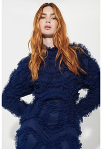 Marinblå klänning med vågig struktur, långa ärmar och hög halsringning. Utrustad med lager av volanger hela vägen, vilket ger dimension och detalj.