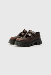 Scarpe in pelle marrone con ritagli e due fibbie regolabili in argento. Suole nere spesse con una trama rigata. Accenti di design wingtip.