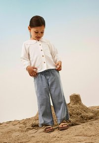 Camisa blanca de manga larga con cuello de encaje y botones de madera, combinada con pantalones vaqueros azules holgados y sandalias marrones sobre la arena.