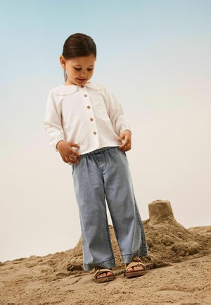 Vit långärmad skjorta med spetskrage och träknappar, matchad med lösa blå denimbyxor och bruna sandaler på sand.