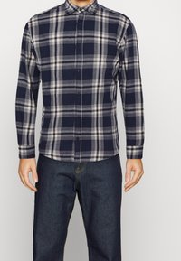 Camicia a quadri blu navy e grigia con bottoni, realizzata in un tessuto morbido, caratterizzata da maniche lunghe e colletto classico. Abbinata a jeans di denim scuri.