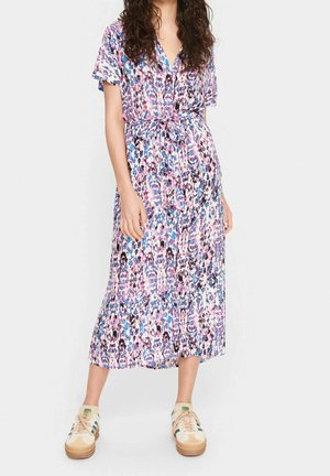 Mujer de pie con un vestido midi de manga corta y cinturón, con un patrón abstracto en rosa, azul y negro, y zapatillas de color claro.