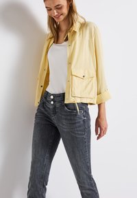Gul lättviktsjacka med silverfärgade tryckknappar, ett bröstfack; kombineras med mörkgrå jeans med två knappar och framfickor.