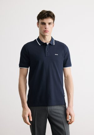 Mladý muž v tmavě modrém polokošili s bílým lemováním a logem "BOSS", v kombinaci se šedými kalhotami, stojící před jednobarevným pozadím.