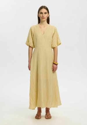 Kobieta stojąca, ubrana w długą, teksturowaną jasnżółtą sukienkę z bufiastymi rękawami, dekoltem w serek, sandałami i dwoma bransoletkami na prawym nadgarstku.