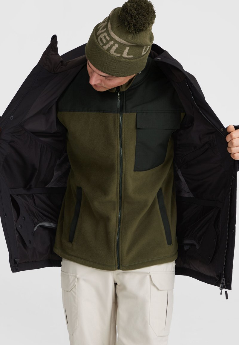 Zielona i czarna polarowa kurtka z zamkiem błyskawicznym z przodu, kieszenią na piersi i kieszeniami bocznymi, noszona rozpięta na jasnobeżowej warstwie bazowej i spodniach.