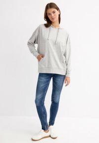 Sudadera con capucha de un gris claro, con un bolsillo delantero, de corte holgado y mangas largas, combinada con jeans ajustados azules y zapatillas blancas con suela color tostado.