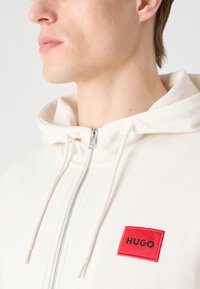 Felpa bianca con zip che presenta una toppa rettangolare rossa con "HUGO" in nero. Tessuto morbido con cappuccio regolabile e chiusura con zip.