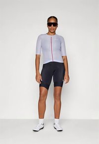 Γυναίκα που στέκεται φορώντας ένα ανοιχτό γκρι κοντομάνικο ποδηλατικό μπλουζάκι, μαύρες ποδηλατικές βερμούδες, λευκές κάλτσες και παπούτσια, και μεγάλες μαύρες αθλητικές γυαλιά ηλίου.