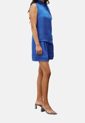 Top azul real sin mangas con cuello de lazo, combinado con pantalones cortos a juego. La modelo lleva tacones plateados con tiras y diseño de tacón grueso.